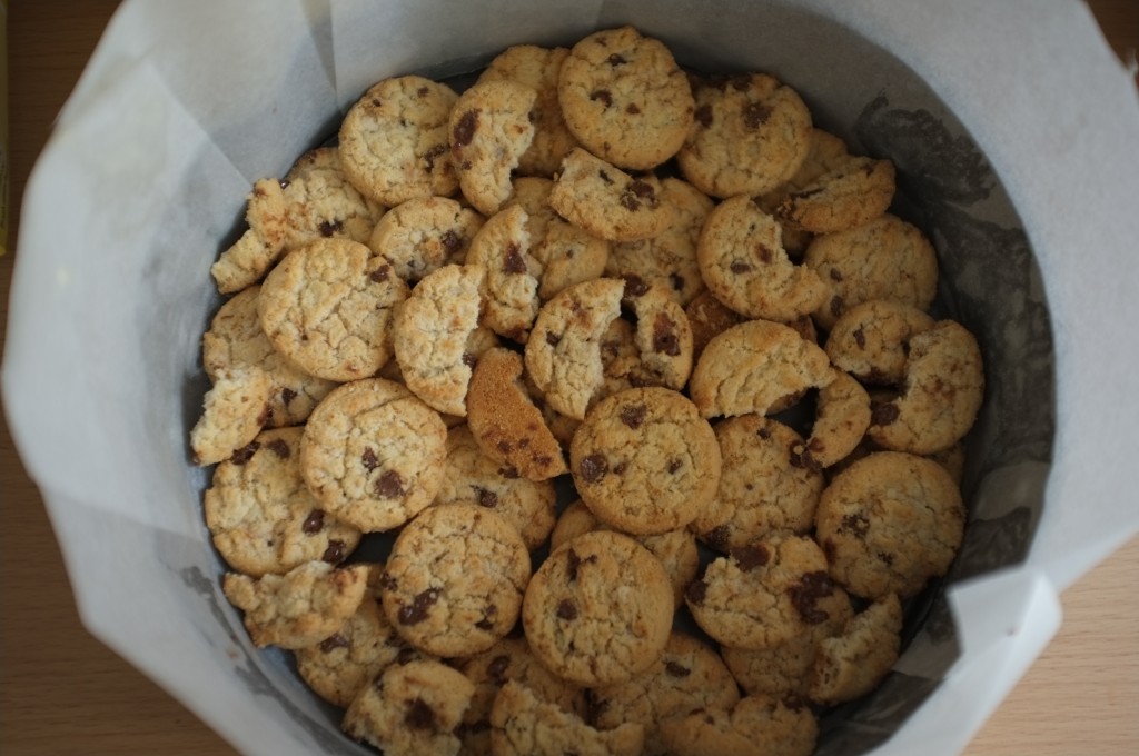 Les cookies sans gluten sont déposés au fond du moule formé un fond de gâteau bien épais. On apperçoit les hauts bords du papier cuisson. 