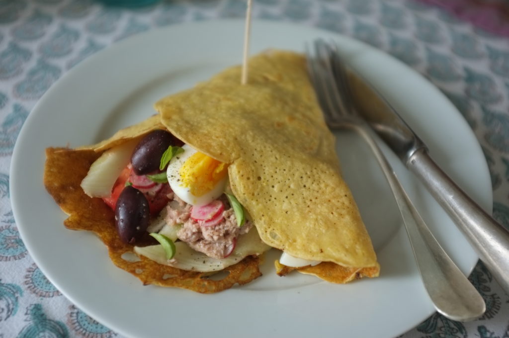 la crêpe Socca sans gluten à la farine de pois chiche, garnie de crudité, thon, olives et oeufs durs. La Socca est présentée dans une assiette blanche.